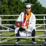 man wears white graduation gown and holds up diploma on bleachers during daytime near grass and trees