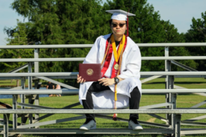 man wearing white graduation gown and holds up diploma on bleachers during daytime near grass and trees