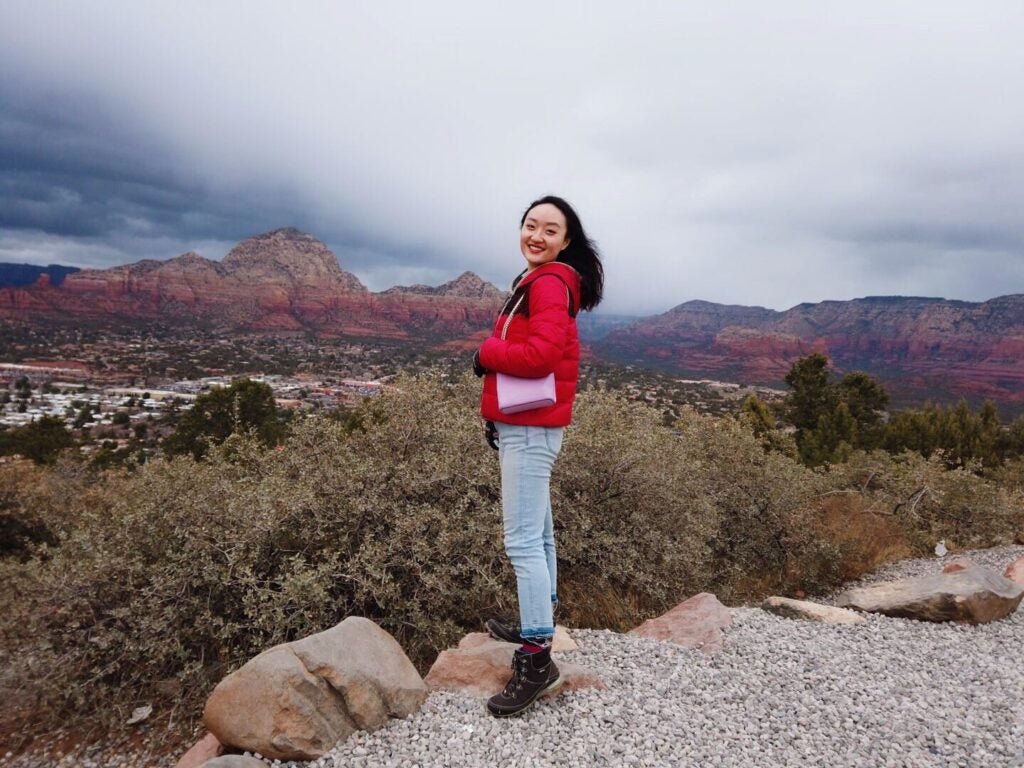 Yuwan from South Korea smiles in a red jacket with a light pink purse while standing in front of an expansive mountain range in Sedona, AZ
