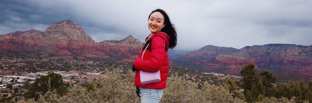 Yuwan from South Korea smiles in a red jacket with a light pink purse while standing in front of an expansive mountain range in Sedona, AZ