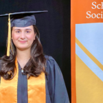 woman in graduation cap and gown with orange sash stands next to orange banner