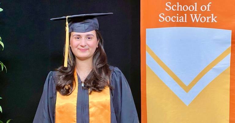 woman in graduation cap and gown with orange sash stands next to orange banner