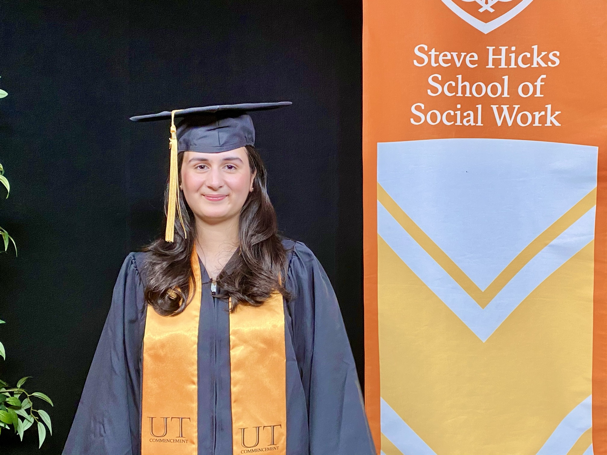 woman in graduation cap and gown with orange sash stands next to orange banner 