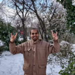 Kumar stands in a brown jacket in front of a snow pile, his hands are in the "hook 'em" position.