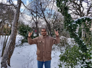 Kumar stands in a brown jacket in front of a snow pile, his hands are in the "hook 'em" position.