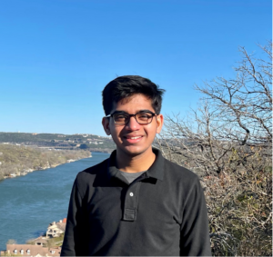 Goenka wears a black collared shirt and stands in front of the blue sky and river view from Mount Bonnell.