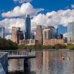 Bikers and Kayakers enjoy Lady Bird Lake and trail in front of Austin city view on a blue day.
