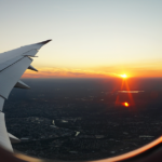 View looks through window, out at the plane wing, the sunset, and urban landscape below.