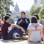 Students sit in a circle on the South Mall with the Texas capitol behind them