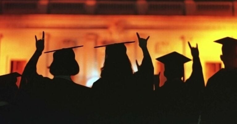 ISSS grads look towards the illuminated UT main building while showing the hook'em sign in their graduation gowns