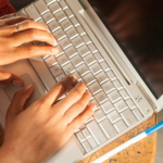 Close up image of someone typing on a laptop computer with two hands. A blue pen sits next to the portable computer.