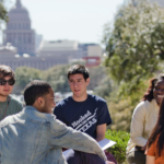 A group of students sit in a circle and have a light-hearted conversation on the UT campus. The Texas State Capitol is in the background.