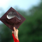A student holds up a black graduation cap with a red tassel outside