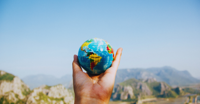 Image of a single hand holding a small globe with a mountain landscape in the background.