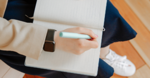 Student writing in a journal. Student is wearing blue jeans and tennis shoes, a black watch, a cream colored shirt.