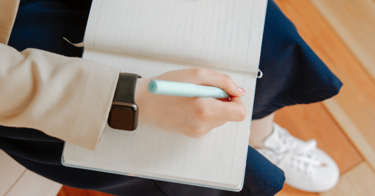 Student writing in a journal. Student is wearing blue jeans and tennis shoes, a black watch, a cream colored shirt.