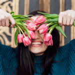A woman in a blue sweater holds up pink tulips in both hands and smiles large at the camera.
