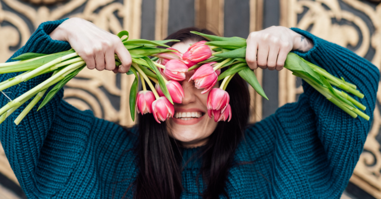 A woman in a blue sweater holds up pink tulips in both hands and smiles large at the camera.