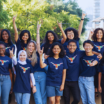 A large group of university students stand outside on campus holding up their arms and making the "longhorn" hand symbol with their middle fingers down and their thumb and pinky finger out.