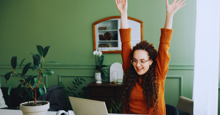 A woman in an orange shirt holds both hands over her head while working on a computer indicating that something positive happened.