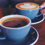 Two cups of coffee in a blue mug and dark blue saucer sit on top of a wooden table