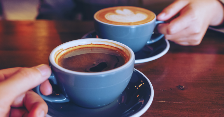 Two cups of coffee in a blue mug and dark blue saucer sit on top of a wooden table