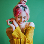 girl in a yellow top, pink hair, and green background smiles away from the camera