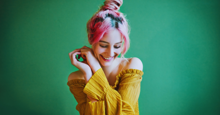 girl in a yellow top, pink hair, and green background smiles away from the camera