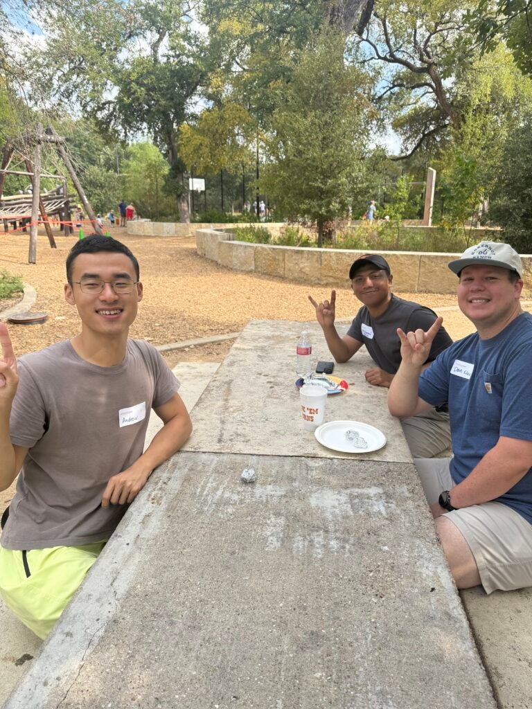 Three male students sit together along a long table at a park