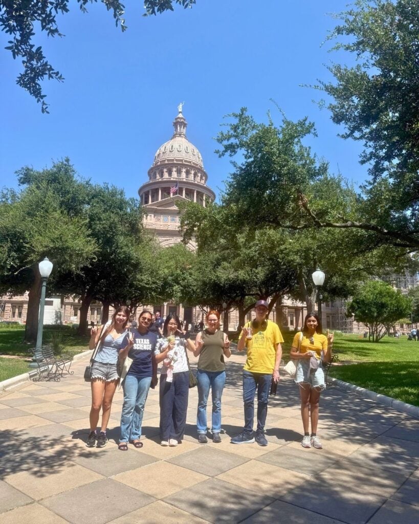 A group of seven smiling people standing in front of a historic capitol building surrounded by trees, under a clear blue sky, conveying a cheerful vibe.