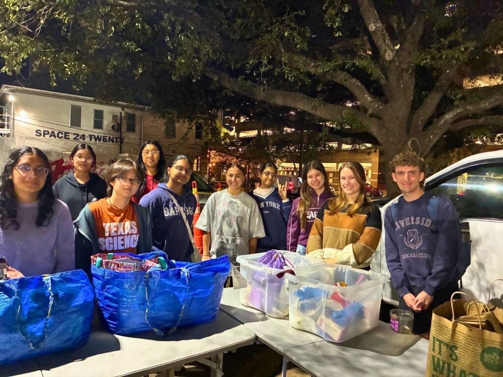 A group of diverse young people stands behind tables filled with bags and bins under a large tree at night, smiling and conveying a sense of community.
