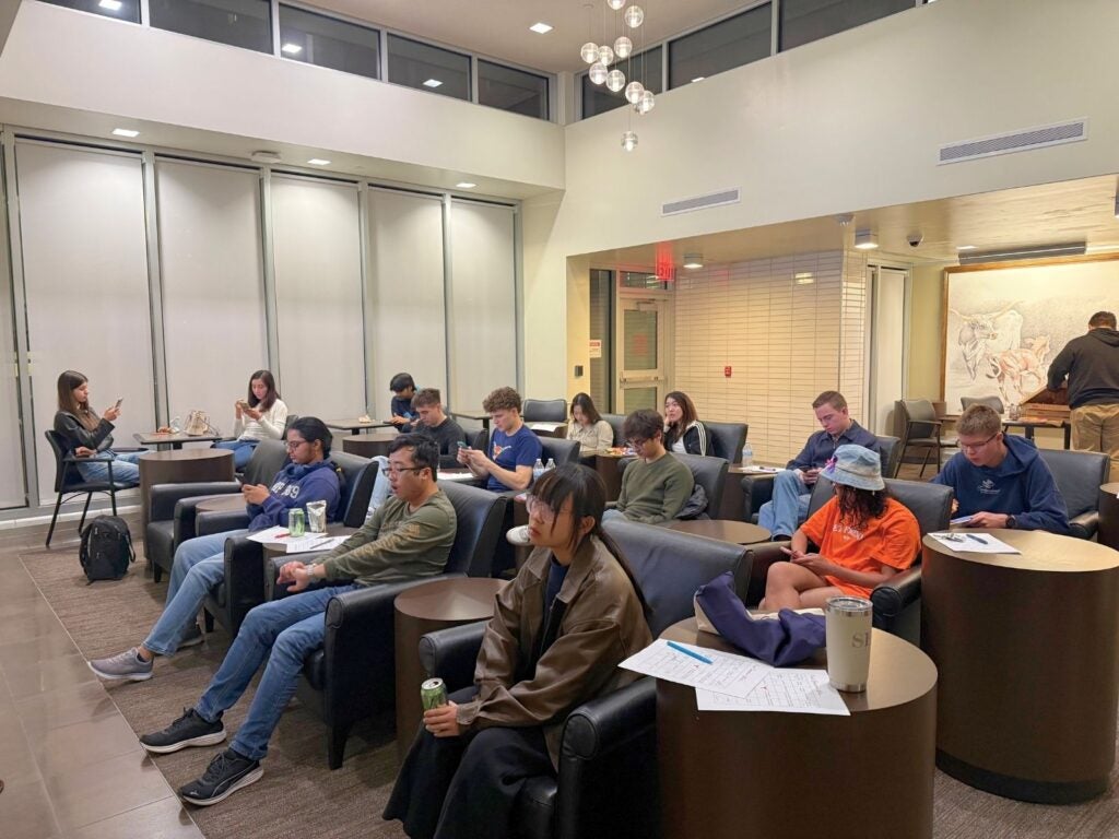A group of people sit in a modern lounge, focused on their phones. The room is bright with natural and artificial light, creating a calm atmosphere.