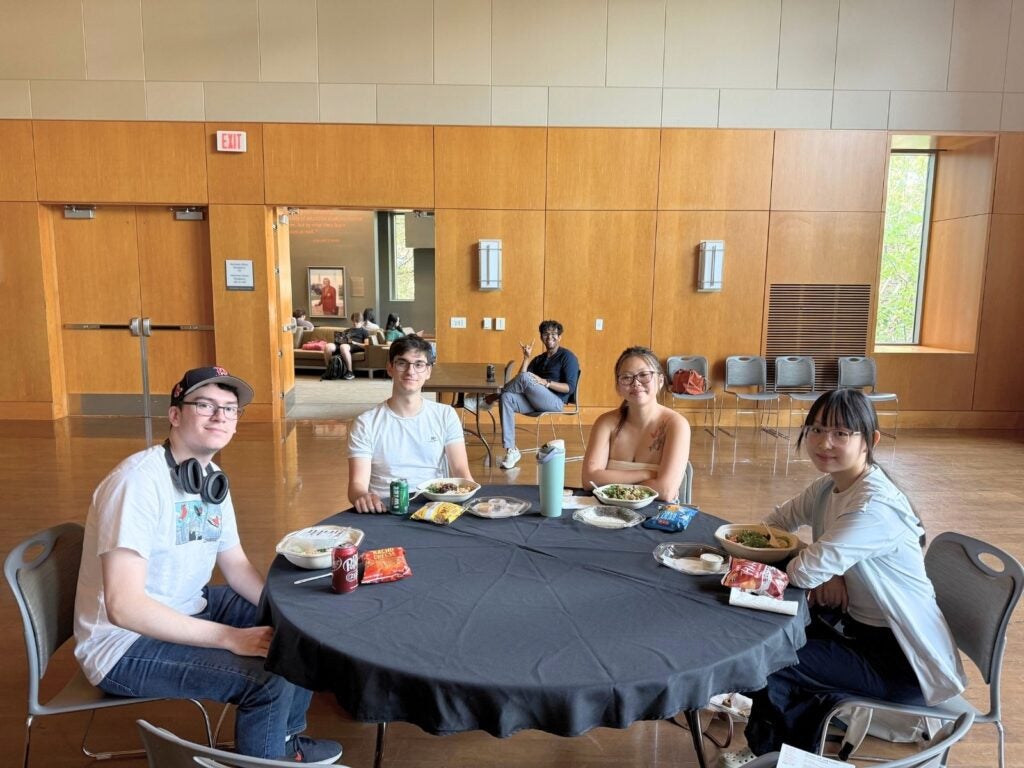 Four people sit at a round table with various lunch items. A person is in the background. The setting is a bright room with wooden walls.
