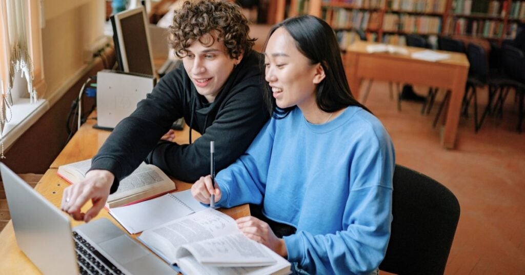 Two students, a young man and woman, smile while working together at a library table. They are focused on a laptop and books, creating a collaborative, studious mood.