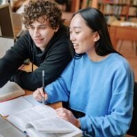 Two students, a young man and woman, smile while working together at a library table. They are focused on a laptop and books, creating a collaborative, studious mood.