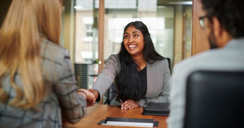 A woman with long dark hair smiles warmly, shaking hands with a blonde woman across a table.