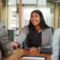 A woman with long dark hair smiles warmly, shaking hands with a blonde woman across a table.