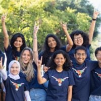 A diverse group of people, wearing navy blue shirts with a multicolored bull logo, smile and raise their hands, forming a welcoming, joyful scene outdoors.