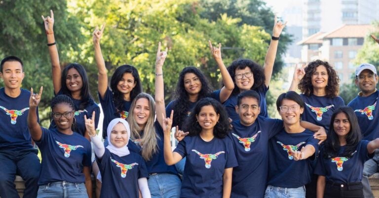 A diverse group of people, wearing navy blue Texas Global shirts with UT longhorn imagery, smile and raise their hands, forming a welcoming, joyful scene outdoors.
