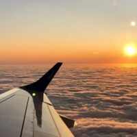Airplane wing in foreground against a backdrop of dense, fluffy clouds at sunset; warm orange and golden sky conveying peace and serenity.