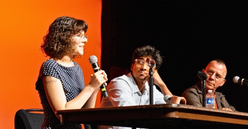 A woman speaks into a microphone at a panel discussion. She is seated next to two men who are attentively listening. The background is a warm orange color.