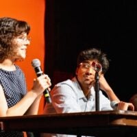 A woman speaks into a microphone at a panel discussion. She is seated next to two men who are attentively listening. The background is a warm orange color.