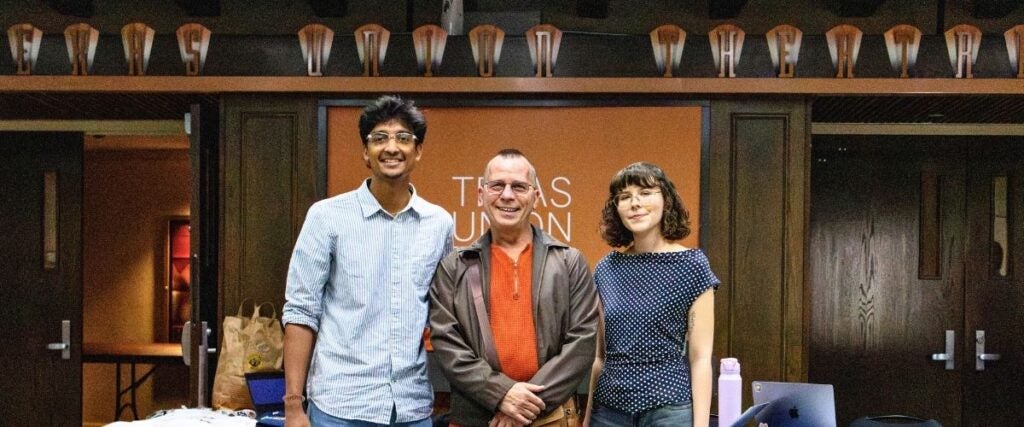Two men and a woman stand smiling in front of a "Texas Union" sign. They appear relaxed and friendly. A table with tech items is partially visible.