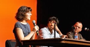 A woman speaks into a microphone at a panel discussion. She is seated next to two men who are attentively listening. The background is a warm orange color.