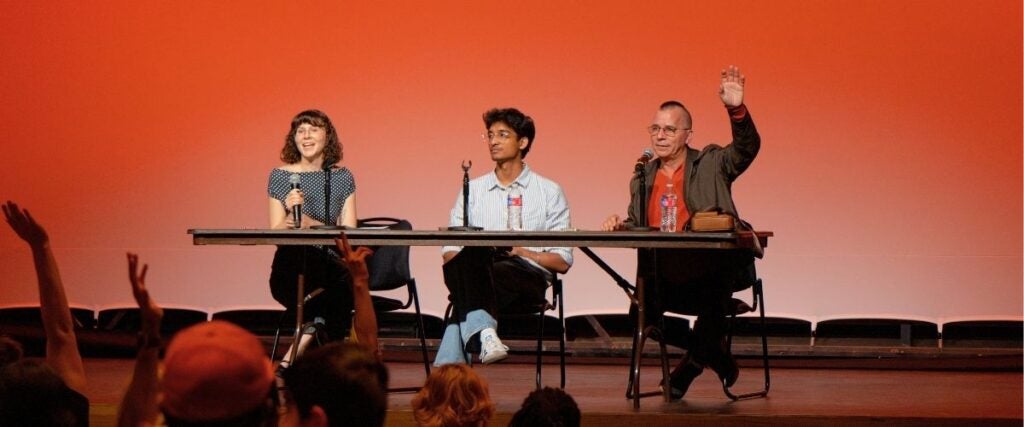 A woman and two men sit at a table on stage with microphones. The woman on the left smiles, the man in the middle looks attentive, and the man one on the right raises a hand. The background is a warm orange gradient. Audience members raise their hands. The atmosphere is engaging and lively.