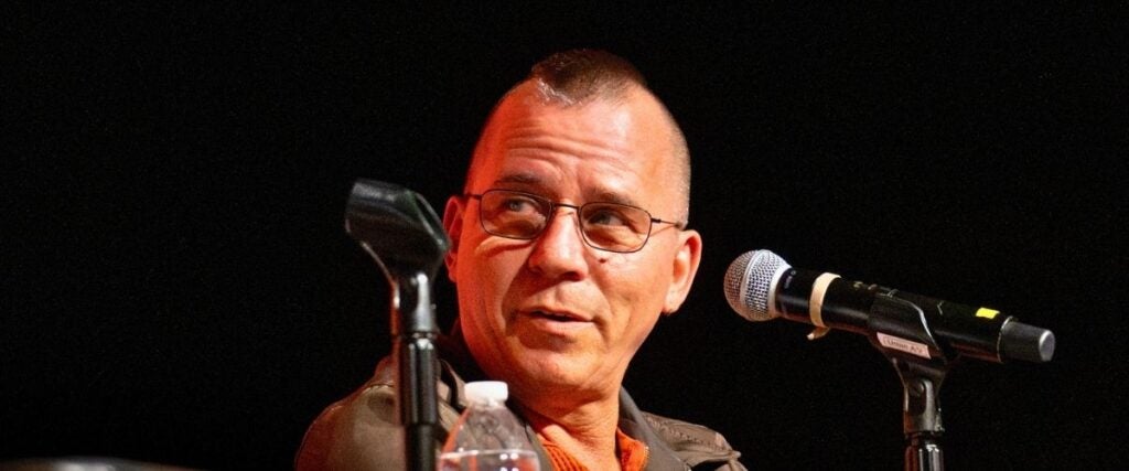 Man with glasses speaks into a microphone on stage. He wears an orange shirt and jacket, with a water bottle and another microphone nearby.