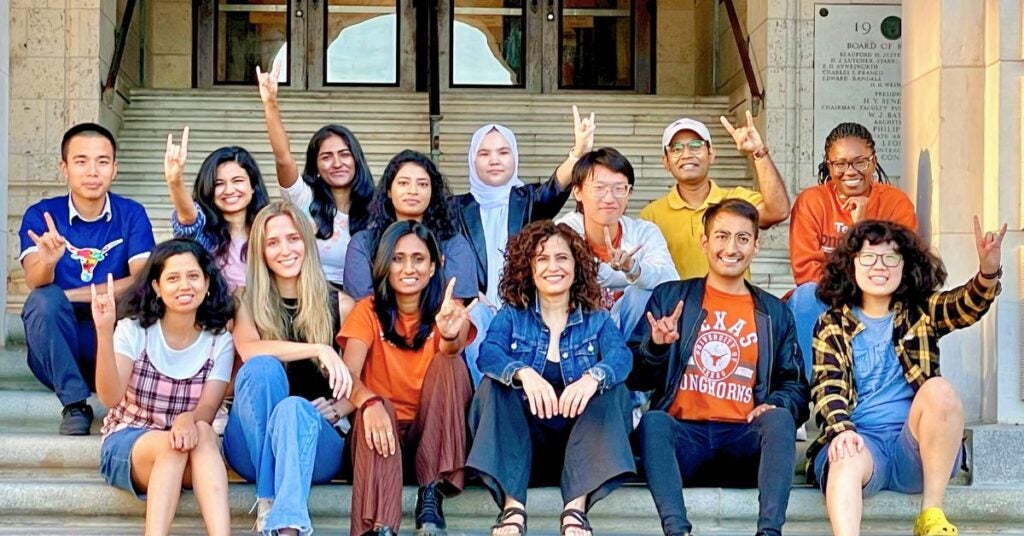 A diverse group of fifteen people sits on steps, smiling and making hand gestures. They convey a joyful and united atmosphere. The background features a building entrance.