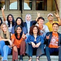 A diverse group of fifteen people sits on steps, smiling and making hand gestures. They convey a joyful and united atmosphere. The background features a building entrance.