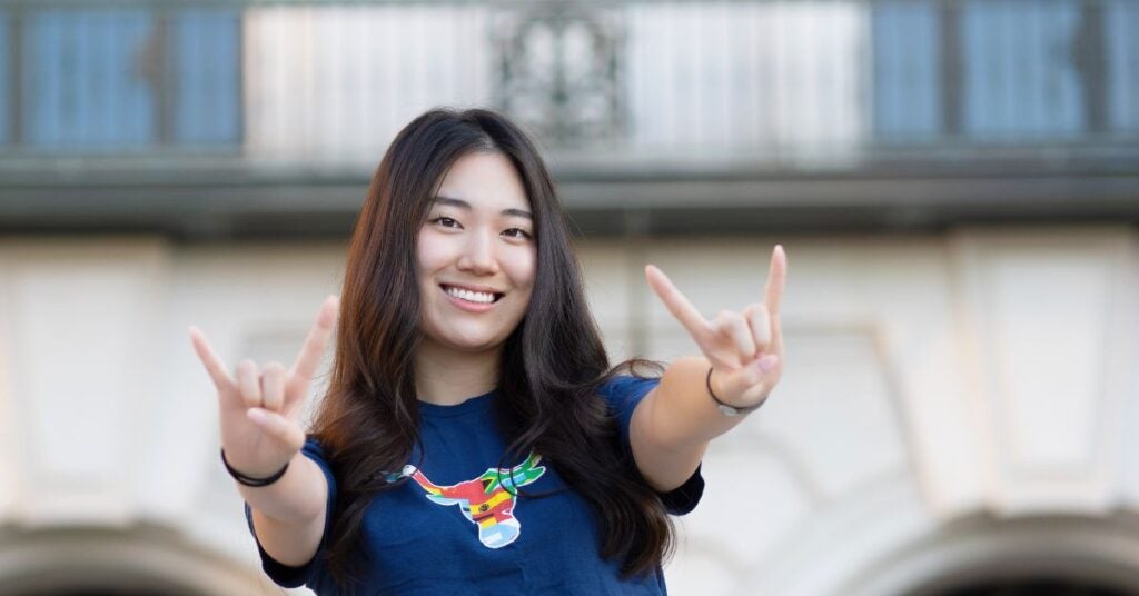 A young woman wearing a navy blue shirt with a colorful bull graphic making a Texas longhorns hand signs outdoors.