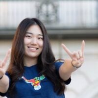 A young woman wearing a navy blue shirt with a colorful bull graphic making a Texas longhorns hand signs outdoors.